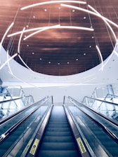 A modern escalator ascends towards a ceiling adorned with curved, illuminated light fixtures. The ceiling features a circular wooden panel that contrasts with the sleek glass and metal elements of the escalator.