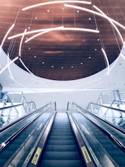 A modern escalator ascends towards a ceiling adorned with curved, illuminated light fixtures. The ceiling features a circular wooden panel that contrasts with the sleek glass and metal elements of the escalator.