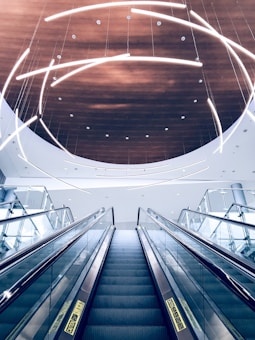 A modern escalator ascends towards a ceiling adorned with curved, illuminated light fixtures. The ceiling features a circular wooden panel that contrasts with the sleek glass and metal elements of the escalator.