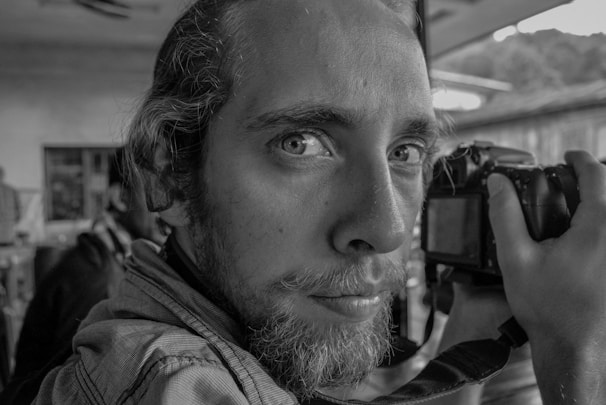 A close-up black and white portrait captures the intense gaze of a person holding a camera. The background is slightly blurred, suggesting an indoor setting with natural light coming through a window.