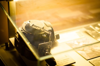 A vintage camera or measuring device is displayed inside a glass case with warm lighting. Accompanying it are several pieces of paper, possibly informational cards, and photographs.
