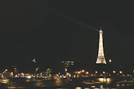 The Eiffel Tower illuminated at night with sparkling lights, viewed from the Seine riverbank in Paris.