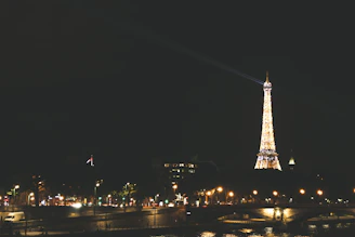 The Eiffel Tower illuminated at night with sparkling lights, viewed from the Seine riverbank in Paris.