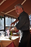 A man with gray hair is using a corkscrew to open a bottle of wine in a warmly lit restaurant setting. He is wearing a dark, striped shirt and a black apron. The table is covered with an orange tablecloth, and a wine bottle and glass are visible. Sunlight is streaming in through the windows.