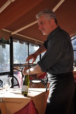 A man with gray hair is using a corkscrew to open a bottle of wine in a warmly lit restaurant setting. He is wearing a dark, striped shirt and a black apron. The table is covered with an orange tablecloth, and a wine bottle and glass are visible. Sunlight is streaming in through the windows.