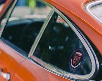 A close-up view of an orange car door featuring a window with a smooth chrome trim. Affixed to the window is a circular decal with 'Porsche 1976 World Champion' displayed prominently in red and black colors. The background is blurred, bringing attention to the car and decal.