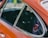 A close-up view of an orange car door featuring a window with a smooth chrome trim. Affixed to the window is a circular decal with 'Porsche 1976 World Champion' displayed prominently in red and black colors. The background is blurred, bringing attention to the car and decal.