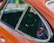 A close-up view of an orange car door featuring a window with a smooth chrome trim. Affixed to the window is a circular decal with 'Porsche 1976 World Champion' displayed prominently in red and black colors. The background is blurred, bringing attention to the car and decal.