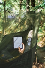 A person is lying in a hammock made of green netting, surrounded by trees and foliage. They are holding a tablet device displaying the text 'YBOOK', suggesting reading or browsing.
