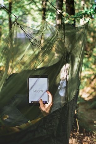 A person is lying in a hammock made of green netting, surrounded by trees and foliage. They are holding a tablet device displaying the text 'YBOOK', suggesting reading or browsing.