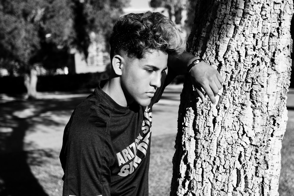 gray scale photo of man leaning on tree trunk