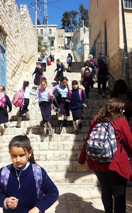 A group of children in school uniforms descends a stone staircase in an urban setting. The school uniforms consist of blue shirts and navy skirts, and many are carrying colorful backpacks. Adults accompany them, some wearing different attire. The scene is outdoors, with buildings lining the stairs and a clear blue sky above.