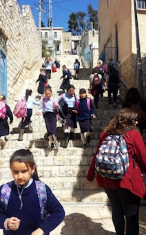 A group of children in school uniforms descends a stone staircase in an urban setting. The school uniforms consist of blue shirts and navy skirts, and many are carrying colorful backpacks. Adults accompany them, some wearing different attire. The scene is outdoors, with buildings lining the stairs and a clear blue sky above.