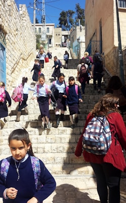 A group of children in school uniforms descends a stone staircase in an urban setting. The school uniforms consist of blue shirts and navy skirts, and many are carrying colorful backpacks. Adults accompany them, some wearing different attire. The scene is outdoors, with buildings lining the stairs and a clear blue sky above.