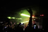 A lively concert scene with a crowd of people raising their hands and capturing the moment with their smartphones. Bright green and yellow lights illuminate the stage, where a DJ is performing. The mood is energetic and vibrant.