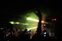 A lively concert scene with a crowd of people raising their hands and capturing the moment with their smartphones. Bright green and yellow lights illuminate the stage, where a DJ is performing. The mood is energetic and vibrant.