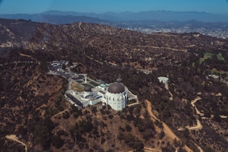 Aerial view of a large, white observatory with distinctive domes situated in a hilly, arid landscape. The surrounding terrain is dotted with dry vegetation and interspersed with winding dirt paths. A sprawling cityscape is visible in the distance, nestled among mountains.