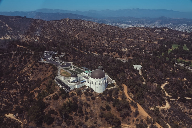 Aerial view of a large, white observatory with distinctive domes situated in a hilly, arid landscape. The surrounding terrain is dotted with dry vegetation and interspersed with winding dirt paths. A sprawling cityscape is visible in the distance, nestled among mountains.