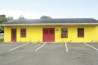A one-story building with a vivid yellow facade and contrasting red doors and window frames stands in front of an empty parking lot. The roof is flat and covered in dark shingles. Sparse vegetation and cracks are visible on the asphalt, and trees are in the background under a partly cloudy sky.