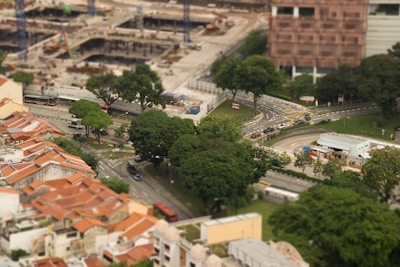 A view of an urban area with red-roofed residential buildings on the left, surrounded by lush green trees. A construction site occupies the background with cranes and unfinished structures. Several roads with moving vehicles intersect the area, and a modern building stands on the right.