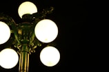 A vintage street lamp with five illuminated globes stands against a dark background. The lamp's structure is ornate, with a greenish tint from the reflection of the light.
