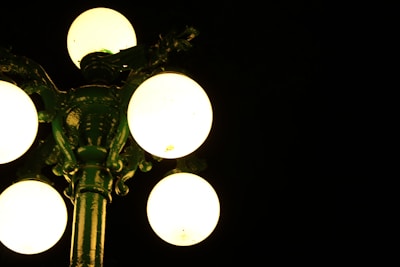 A vintage street lamp with five illuminated globes stands against a dark background. The lamp's structure is ornate, with a greenish tint from the reflection of the light.