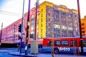 A vibrant urban street scene featuring a red Metro bus in the foreground and an industrial-looking building in the background with colorful geometric artwork on its facade. The area is marked by power lines, street signs, crosswalks, and a chain-link fence, conveying a busy city atmosphere.