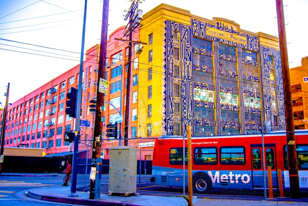 A vibrant urban street scene featuring a red Metro bus in the foreground and an industrial-looking building in the background with colorful geometric artwork on its facade. The area is marked by power lines, street signs, crosswalks, and a chain-link fence, conveying a busy city atmosphere.