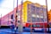 A vibrant urban street scene featuring a red Metro bus in the foreground and an industrial-looking building in the background with colorful geometric artwork on its facade. The area is marked by power lines, street signs, crosswalks, and a chain-link fence, conveying a busy city atmosphere.