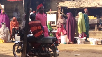 A group of people are gathered in an outdoor marketplace. Many are wearing colorful, traditional clothing, including vibrant hijabs and caps. They appear to be engaged in conversation and buying or selling goods, with several plastic containers visible. A motorcycle with a person seated on it is in the foreground.