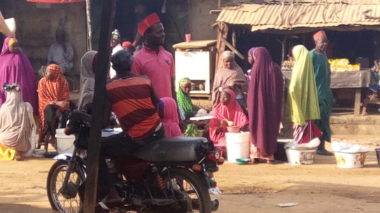 A group of people are gathered in an outdoor marketplace. Many are wearing colorful, traditional clothing, including vibrant hijabs and caps. They appear to be engaged in conversation and buying or selling goods, with several plastic containers visible. A motorcycle with a person seated on it is in the foreground.