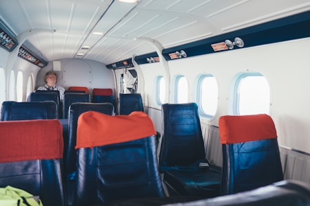 The interior of a small aircraft with several rows of empty black seats adorned with red headrest covers. A single passenger is seated towards the back, wearing a striped shirt. The cabin has a clean, functional design with overhead lights and windows along the side, allowing natural light to filter in.