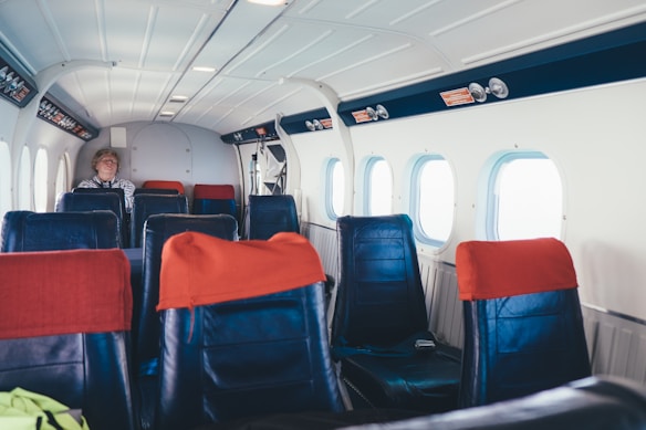 The interior of a small aircraft with several rows of empty black seats adorned with red headrest covers. A single passenger is seated towards the back, wearing a striped shirt. The cabin has a clean, functional design with overhead lights and windows along the side, allowing natural light to filter in.