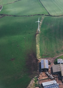 Aerial view of a green agricultural field featuring a wind turbine. Below the turbine, a farm with buildings equipped with solar panels is visible. There are cows grazing near the farm building and scattered in the field.