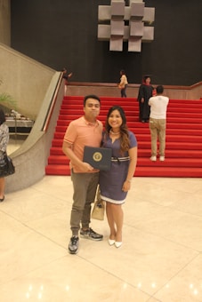A man and a woman are standing at the bottom of a grand staircase with red carpeted steps. The man is holding a certificate, suggesting a special occasion or celebration. In the background, a few other people can be seen on the stairs, including someone in a graduation gown and cap. The setting appears to be an elegant indoor space, possibly a hall or auditorium, with modern decor.