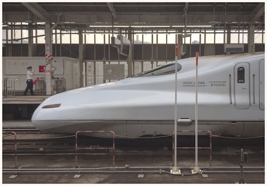 A sleek, modern high-speed train at a station platform. The train is silver with a streamlined design, labeled 'West Japan Kyushu.' A person in uniform is walking along the platform carrying a small red flag, adding a human element to the mechanical scene. The train station is covered, featuring metal beams and industrial architecture, with signage and safety barriers present.