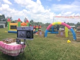 An outdoor setting features colorful inflatable play structures and a decorative balloon arch. A table draped with a pink and white skirt holds electronic equipment, possibly a photo booth setup. In the background, lush greenery and a partly cloudy sky are visible, creating a festive atmosphere.