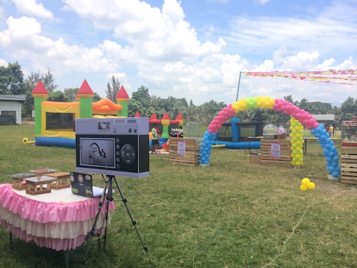 An outdoor setting features colorful inflatable play structures and a decorative balloon arch. A table draped with a pink and white skirt holds electronic equipment, possibly a photo booth setup. In the background, lush greenery and a partly cloudy sky are visible, creating a festive atmosphere.