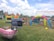 An outdoor setting features colorful inflatable play structures and a decorative balloon arch. A table draped with a pink and white skirt holds electronic equipment, possibly a photo booth setup. In the background, lush greenery and a partly cloudy sky are visible, creating a festive atmosphere.