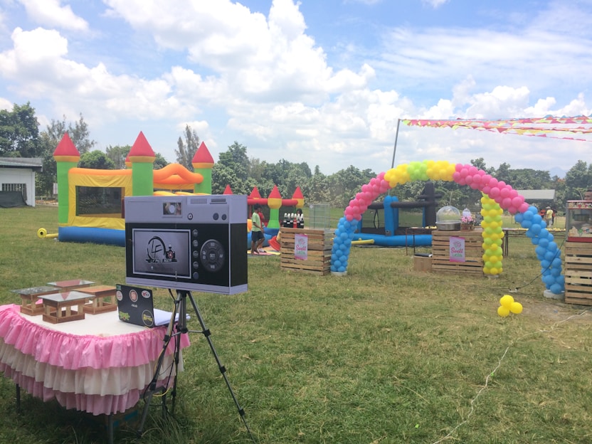 An outdoor setting features colorful inflatable play structures and a decorative balloon arch. A table draped with a pink and white skirt holds electronic equipment, possibly a photo booth setup. In the background, lush greenery and a partly cloudy sky are visible, creating a festive atmosphere.