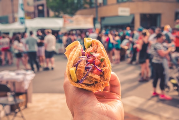 A close-up of a hand holding a taco with vibrant ingredients such as meat, tomatoes, and lettuce. The background is blurred, showing a crowd of people at an outdoor event or festival.