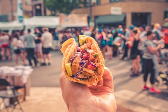 A close-up of a hand holding a taco with vibrant ingredients such as meat, tomatoes, and lettuce. The background is blurred, showing a crowd of people at an outdoor event or festival.