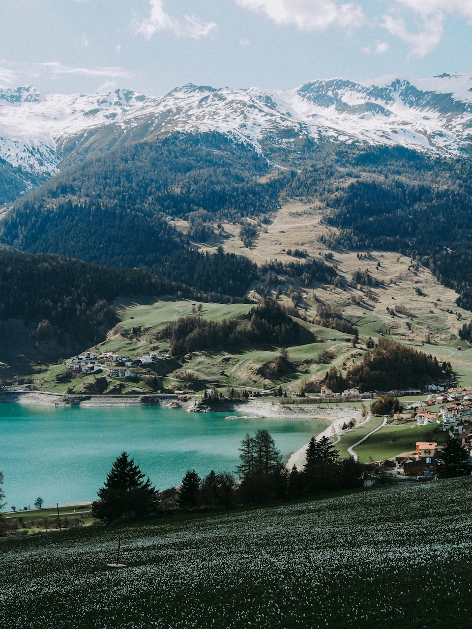 The image depicts a serene mountainous landscape with a large lake at its center, surrounded by lush green fields and trees. The mountains are blanketed in snow, creating a picturesque scene that blends natural beauty with human habitation. The perspective is from the top of one of these mountains, offering a panoramic view of the valley below.