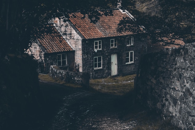 A rustic stone cottage with a weathered red tiled roof is nestled between two stone walls and surrounded by dark, dense foliage. The atmosphere is shadowy and mysterious, with the path leading to the cottage partially obscured by the overhanging branches.