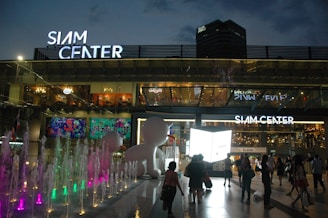 A bustling shopping mall entrance illuminated with bright lights during the evening. People walk towards the entrance, surrounded by colorful water fountains and a large illuminated sign with the name Siam Center. The atmosphere is lively, with vibrant digital displays on the exterior walls.