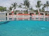 A luxurious poolside with clear blue water reflecting the sky and surrounding palm trees. Several cabanas with red cushions are lined up in the background, providing a sense of relaxation and leisure. The area is adorned with tropical plants, adding to the exotic atmosphere.