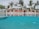 A luxurious poolside with clear blue water reflecting the sky and surrounding palm trees. Several cabanas with red cushions are lined up in the background, providing a sense of relaxation and leisure. The area is adorned with tropical plants, adding to the exotic atmosphere.