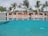 A luxurious poolside with clear blue water reflecting the sky and surrounding palm trees. Several cabanas with red cushions are lined up in the background, providing a sense of relaxation and leisure. The area is adorned with tropical plants, adding to the exotic atmosphere.