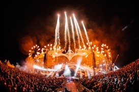 A large outdoor concert at night features bright stage lights and fireworks shooting into the sky, surrounded by a massive crowd of people. The stage has intricate designs with glowing elements, and there is an atmosphere of high energy and excitement as the audience focuses on the performance.