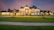 A large, luxurious mansion with intricate architectural details and multiple windows, accented by warm lighting. The house is surrounded by a well-manicured lawn and paved sidewalks, set against a dramatic dusk sky.